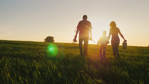 Rear view: A friendly family with a young son is going to plant a tree. Carry a seedling, shovel and watering can. Silhouettes in a beautiful field on a sunset background