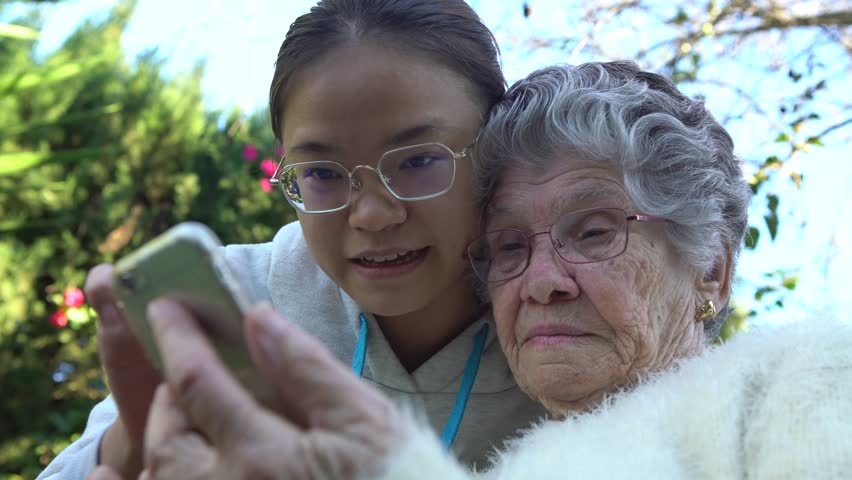 4K Smiling asian granddaughter showing and teaching mobile phone to her grandmother. Happy family, grandchild and grandma using and looking at smart phone in the garden house-Dan