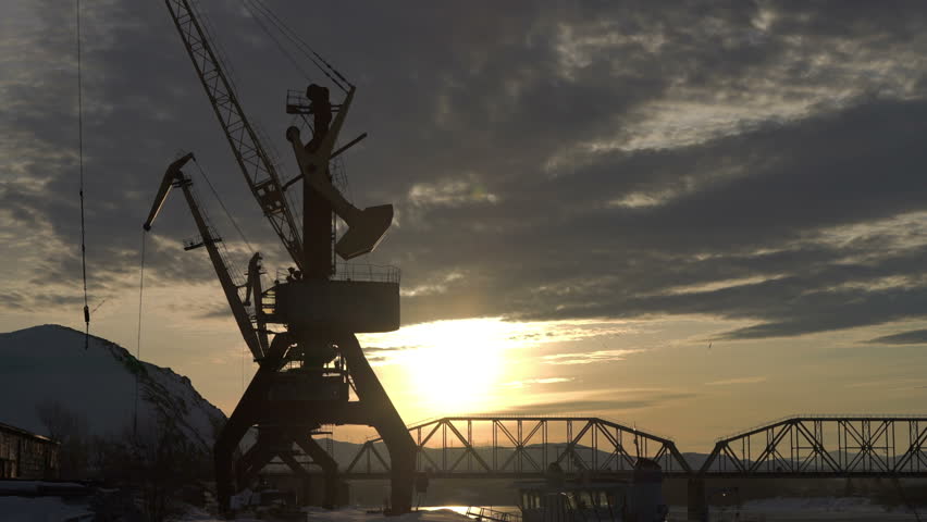 Time lapse of beautiful colorful dusk on a river in winter. Silhouettes of barge, reeds, cranes and railway bridge across the river. Reflections of lights on a water.