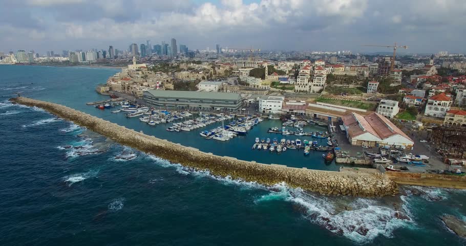 Aerial view high above Ships anchoring at the Jaffa port and people traveling along the coastline in a cloudy day. tel aviv skyline with urban skyscrapers at the background