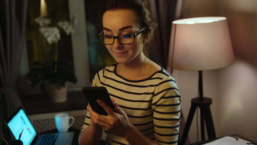 Young business woman in the glasess typing on smart phone at night in office and smilingon window background. Indoor. Close up.