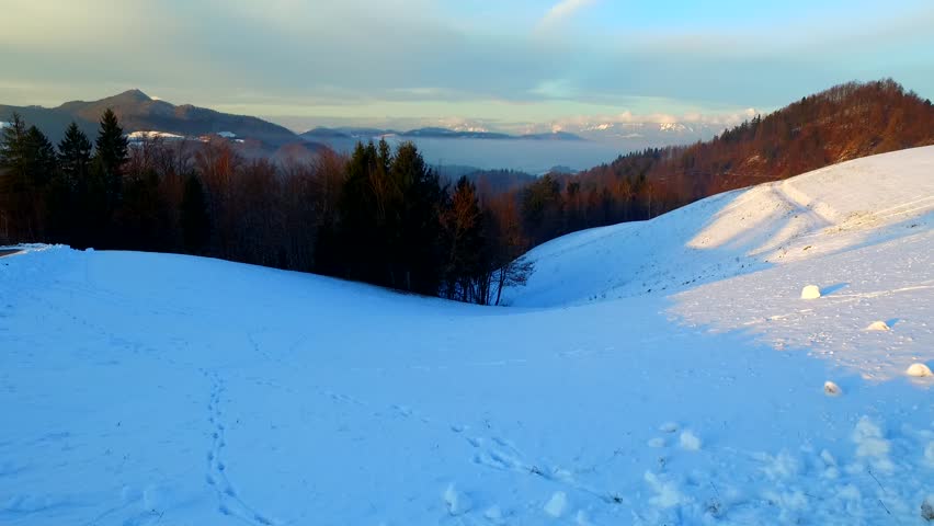 Light setting down under winter landscape with dark spruce forests, distant hills and mountains, covered with some fog and cold mist.