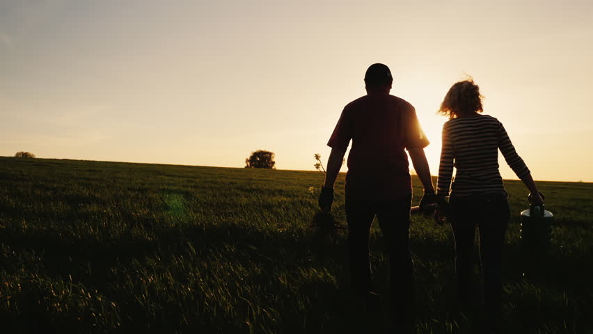 Steadicam shot: Farmers - a man and a woman walking across the field at sunset. Carry a tree seedling, a watering can and a shovel. Concept - work in the garden, volunteering, a new life Royalty-Free Stock Footage #26575091