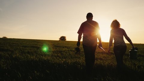 Steadicam shot: Farmers - a man and a woman walking across the field at sunset. Carry a tree seedling, a watering can and a shovel. Concept - work in the garden, volunteering, a new life