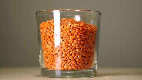 Raw red lentils in transparent glass with white reflection. Cereals pouring into an empty glass on a pastel background, close-up.