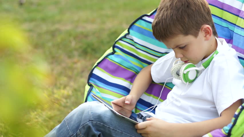 Boy playing with a tablet pc outdoors. Teenager sitting on the chair in jeans
