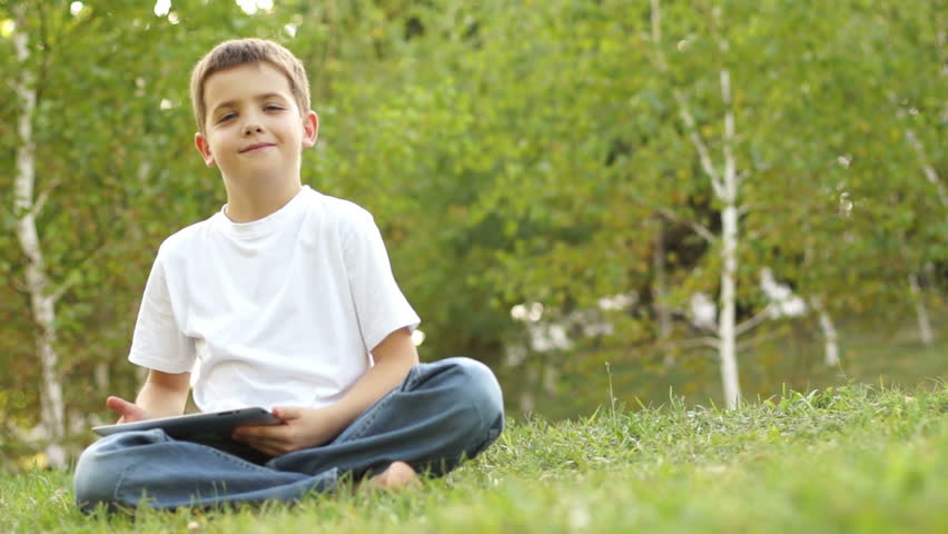 Schoolboy with a tablet pc and sitting on the grass. Thumbs up.