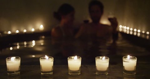 Couple drinking wine on night date in romantic candlelit hot tub spa jacuzzi relaxing in private hotel room resort terrace. Romantic honeymoon getaway travel vacation young adults enjoying holidays.
