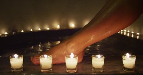 Woman entering hot tub jacuzzi at night for relaxation spa session at resort. Legs closeup, focus on candles. Unrecognizable person enjoying warm water pool therapy at hotel.