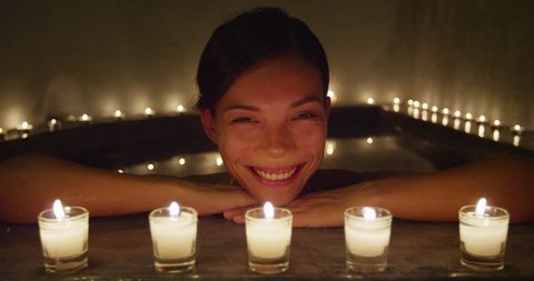 Happy smiling at camera Asian woman relaxing at spa enjoying candlelit night in hot water bath tub jacuzzi in luxury resort private hotel room. Chinese mixed race model face beauty skincare.