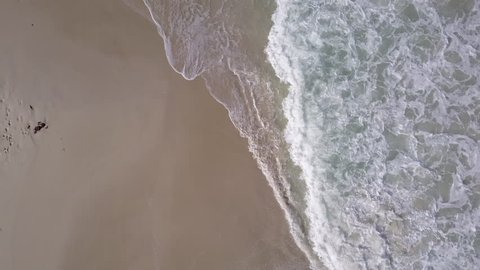 Vertical Aerial view flight along coast, with ocean waves breaking in slow motion at beautiful scenic beach, sand surface as copy space.