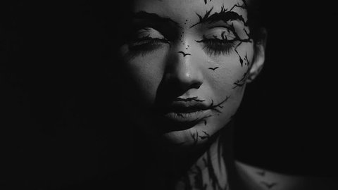 Fashion close-up slow motion portrait of model female with a amazing creative make-up. Painted muah silhouettes of trees and birds. Calm face, halloween. Dark background. Black and White, Contrast.