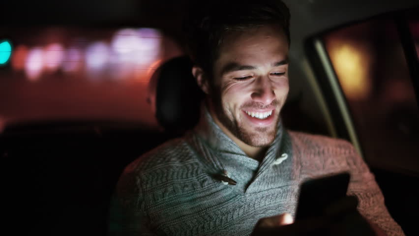 Handsome young man using cell phone in a car. He is texting, checking emails, chats or the news online. Night.
