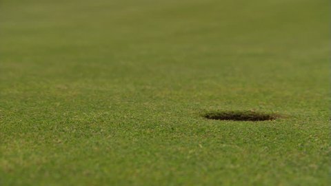 Golf ball rolling from left, stopping on brink of close-up hole at right of frame