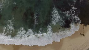 Aerial view looking down, gradually rising high above a beach with waves breaking in as a surfer walks below.