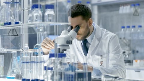 Young Male Research Looks at Sample under Microscope then Looks into Camera and Smiles. He's Sitting in a High-End Modern Laboratory with Beakers, Microscope and Working Monitors Surround Him.
