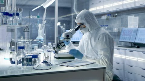 In a Secure High Level Research Laboratory Scientist in a Coverall Examines Petri Dish Under Microscope. Shot on RED EPIC-W 8K Helium Cinema Camera.