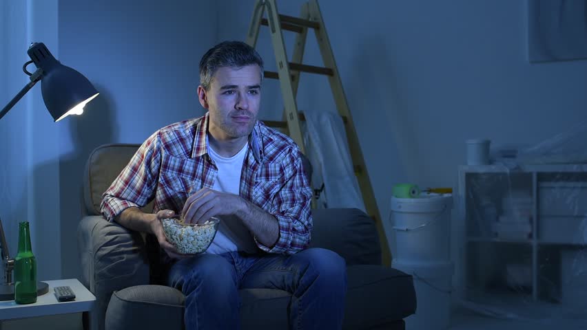 Smiling man relaxing at home at night and watching soccer game on tv, he is cheering and eating popcorn
