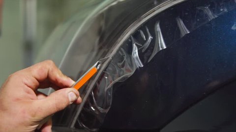 Close up shot of master cutting vinyl film by special knife on the blue car. Expert makes more accurate the pattern for further bonding film on the headlights.