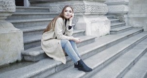 Young stylish female wearing jeans and coat smiling at camera while sitting on stairs of old stone building
