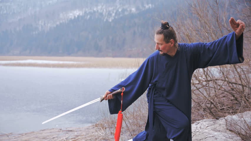 Swordsman warrior in blue kimono posing with sword at lake. Dolly slide medium shot of man standing still while sword pointing towards the lake. Caucasian person with Chinese traditional bun haircut.