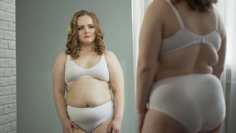 Obese lady in underwear looking at reflection in mirror, ashamed of her fat body, cellulite and stretch marks, appearance insecurities. Fat woman thinking of weightloss program, depression, oversize

