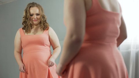 Young woman looking at her mirror reflection with disgust, ashamed of fat body