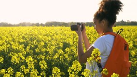 4K video clip of healthy mixed race African American girl teenager female young woman with orange backpack and camera taking photograph in field of rape seed yellow flowers