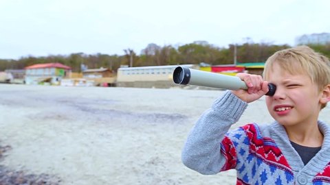 Little child looks through old telescope at something interesting in blue sea. Happy 8 year old kid has fun on beach on vacation in cool spring or autumn season. Real time hd video footage