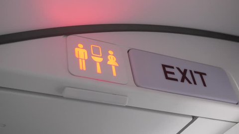 Shot of airplane lavatory sign turning from green (available) to red (occupied). Beside the lavatory sign is an exit sign.