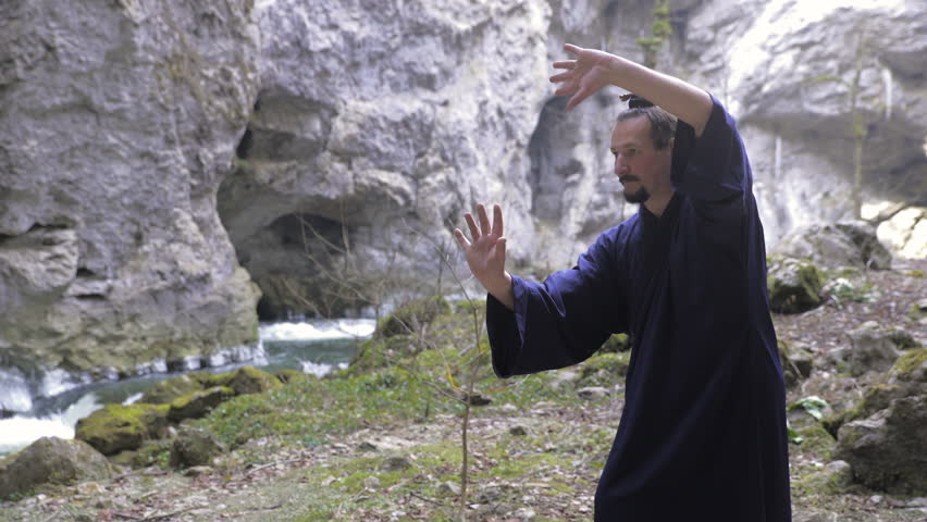 Kung Fu Master Perform Martial Arts Techniques in Cave 4K. Medium jib shot of male person in focus wearing traditional Chinese kimono training Martial Arts. Cave and river in background out of focus.