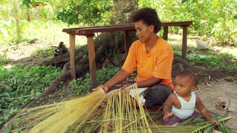 Fijian woman weaving palm fronds with her baby seated nearby