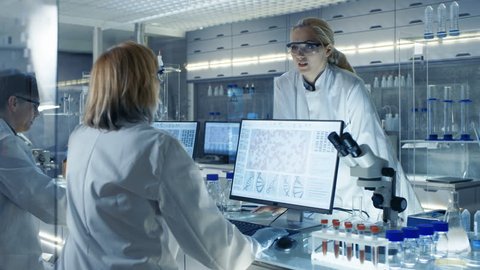 In Modern Laboratory Senior Female Scientist Discusses Work with Young Female Assistant. Laboratory Has Computers, Beakers and Other Technologt for High Tech Scientific Analyses. RED Cinema Camera 8K.