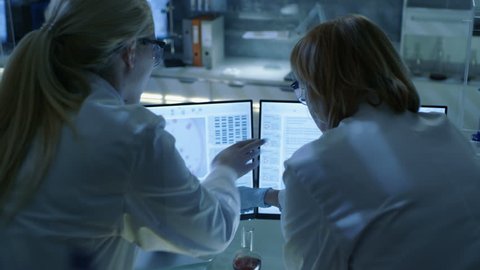 Senior Female Scientist Discusses Scientific Data with Her Laboratory Assistant. They're looking at Two Displays in a Modern Laboratory. Shot on RED EPIC-W 8K Helium Cinema Camera.