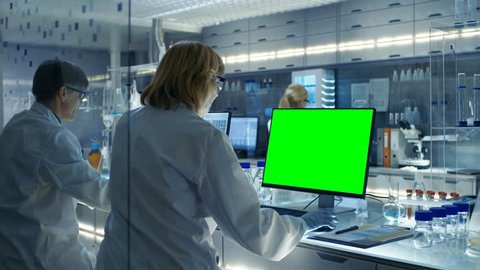 Female and Male Scientists Working on their Computers (Mock-up Green Screen) In Modern Laboratory.Various Shelves with Beakers, Chemicals and Different Technical Equipment is Visible.RED Cinema Camera