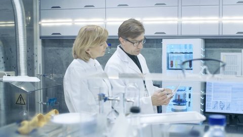 Female and Male Chief Scientists Walking Through Modern Laboratory and Discussing Future Possibilities. Young Woman Assistant Works at Her Desk. Shot on RED EPIC-W 8K Helium Cinema Camera.