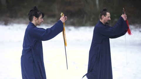 Training Martial Arts Sword Techniques in Snow 4K. Long shot of two person in focus, man and woman in blue kimono train in middle of snow. Medium shot from knees up.
