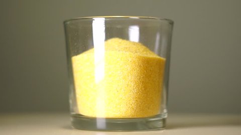 Raw cornmeal polenta in transparent glass with white reflection. Cereals pouring into an empty glass on a pastel background, close-up.