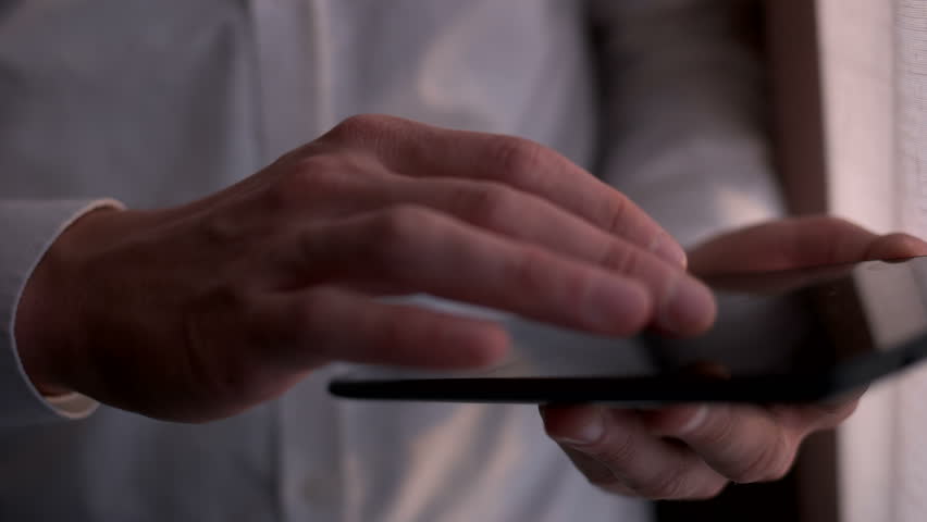 Close up young businessman using tablet computer while standing near window on beautiful sunset. Male hands tap on tablet device toucscreen