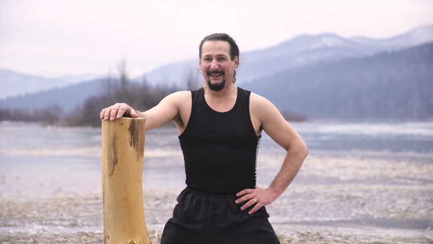 Tired person resting after bodybuilding with lifting log in winter 4K. Long shot of a breathless person in focus holding to tall log after training. Frozen nature in background out of focus.