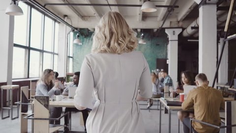 Beautiful blonde female team leader walks through the office, controls the work of employees, gives direction to colleague.