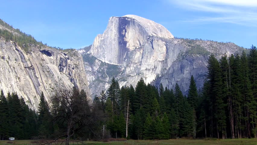 A mountain known as Half Dome rises above a pine forest and green meadow in