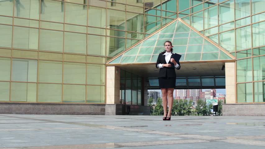 Young confident businesswoman with folder walking near the office building