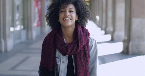 Attractive African girl in scarf with short curly hair posing with hands up and looking cheerfully at camera on background of street.