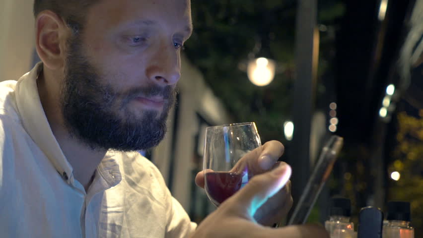 Young man drinking red wine and texting on smartphone in cafe at night

