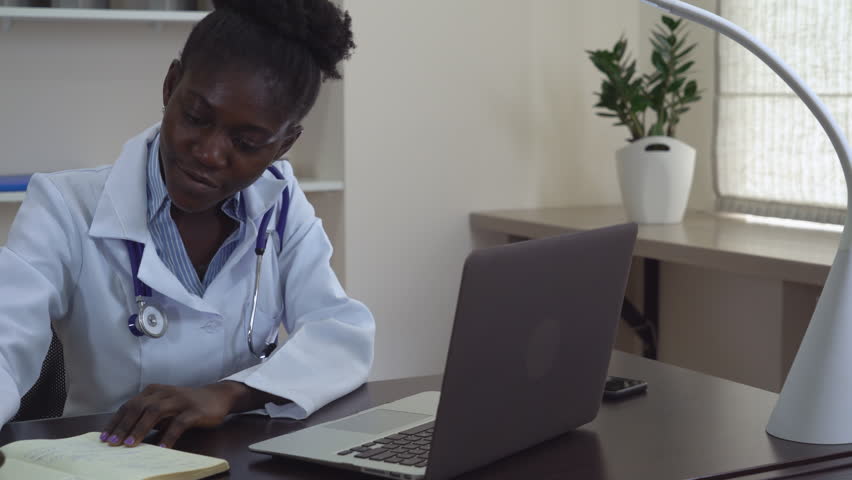 African doctor at working place in clinic. Young professional physician handwriting on notebook. Afro american woman wearing in white coat sitting at the desk with computer Royalty-Free Stock Footage #26850511