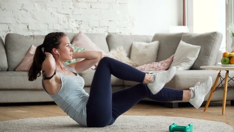 young fit and tone up woman doing fitness workout and sit ups exercises for health using app on mobile phone fixed on hand to listen music in living room at home during sunny day