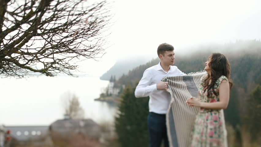 The man is wrapping the lovely girl into the plaid at the background of the mountains.