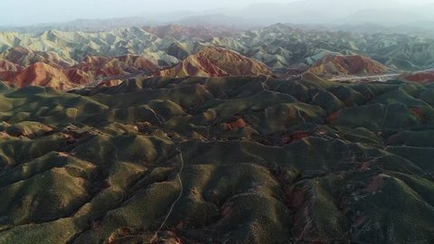 Approaching beautiful rainbow mountains in Zhangye National Geopark, part 2 of a continuous 3 part series. Aerial view on an orange sandstone mountain chain surrounded by green vegetation.