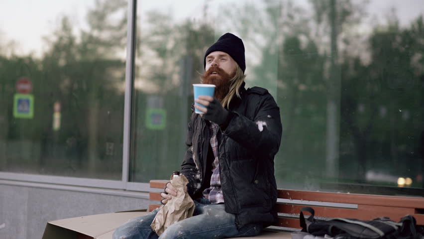 Very drunk homeless man talking to people walking near him and beg for money while sitting on bench at the sidewalk